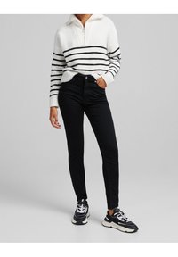 Randig stickad tröja i vitt och svart med dragkedja i kragen, tillsammans med högmidjade svarta jeans och svarta sneakers med vita detaljer.