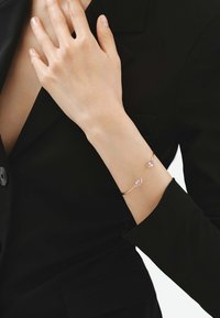 Brazalete de oro con dos piedras ovaladas rosas, exhibido en una mano contra un blazer negro, mostrando un acabado metálico suave.