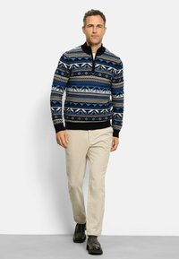 Blau-weiß gemusterter Pullover mit Reißverschlusskragen, der geometrische Designs aufweist. Kombiniert mit hellbeigen Cordhosen und dunklen Schuhen.