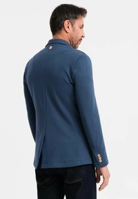 Man i en marinblå kavaj med tre bruna knappar på ärmen och en liten trefärgad tygskylt på baksidan av kragen, stående på sidan.