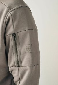Grå sweatshirt med en lynlås-lomme på siden, struktureret stof og et kvadratisk logo-patch på ærmet. Markante syninger.