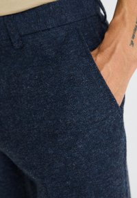 Pantaloni texture blu scuro con un sottile motivo a righe, dotati di tasche laterali e design slim fit. Il tessuto appare morbido e resistente.