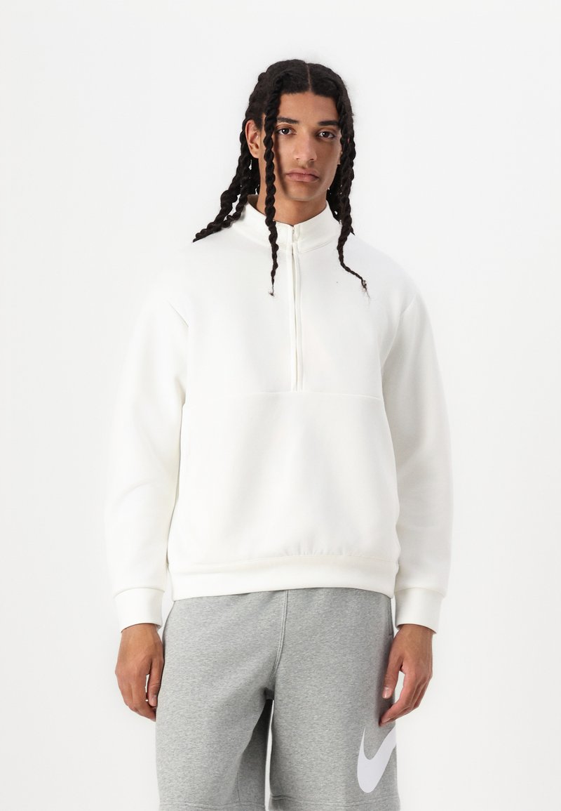 Pullover bianco con colletto alto e mezza zip, caratterizzato da una consistenza liscia e morbida, con una tasca frontale. Indossato con pantaloni della tuta grigi.