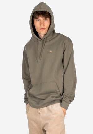 Junger Mann in einem grauen Kapuzensweatshirt mit einem kleinen gestickten Design, beiger Hose, Hände in den Taschen, blickt direkt in die Kamera.