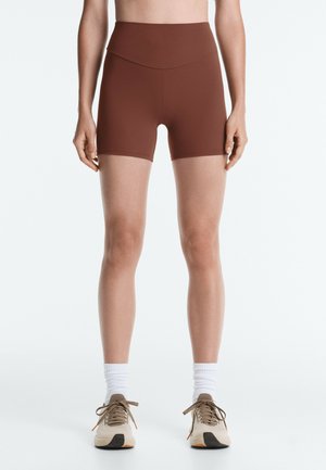 Person trägt hoch taillierte braune Sportshorts, weiße Kniestrümpfe und beige Laufschuhe und steht vor einem schlichten hellen Hintergrund.