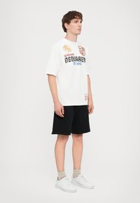 Camiseta blanca de manga corta con gráficos coloridos, texto "DSquared" y parches. Combinada con shorts negros y zapatillas blancas. Material suave.