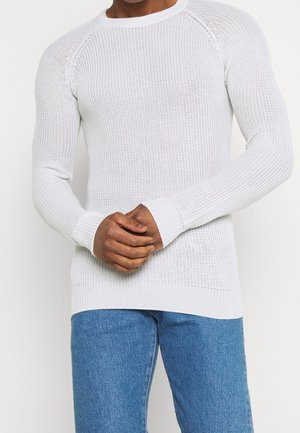 Pull en tricot blanc avec un col rond, des manchettes côtelées et un motif texturé. Le tissu est léger et s'ajuste étroitement au corps.