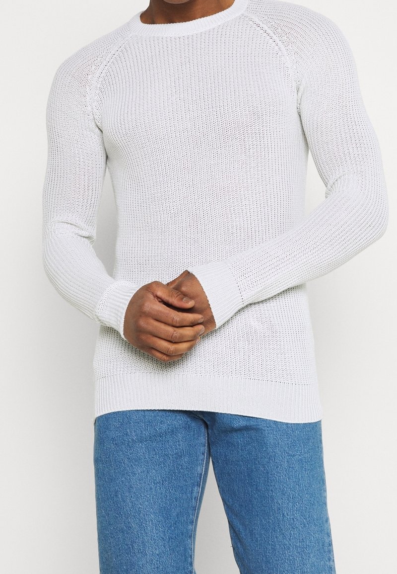 Pull en tricot blanc avec un col rond, des manchettes côtelées et un motif texturé. Le tissu est léger et s'ajuste étroitement au corps.