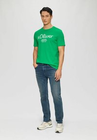Grünes Baumwoll-T-Shirt mit weißem "s.Oliver" Logo, kombiniert mit blauen Jeans und beigen Sneakern. Einfaches Rundhalsdesign.