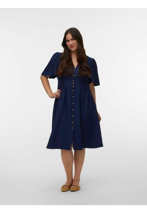 Kvinde med langt mørkt hår iført en marineblå kjole med knapper og korte ærmer samt brune flade sko, stående med den ene hånd på hoften.