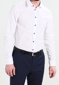 Camicia bianca a maniche lunghe con bottoni blu scuro e un colletto rigido, abbinata a pantaloni blu navy e una cintura nera con fibbia argentata.