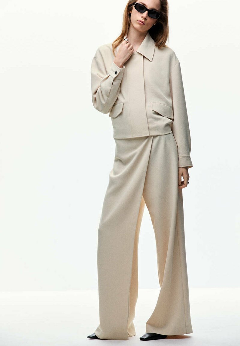 Femme portant une veste beige oversize et un pantalon à jambes larges, chaussures noires et lunettes de soleil foncées, debout devant un fond clair uni.