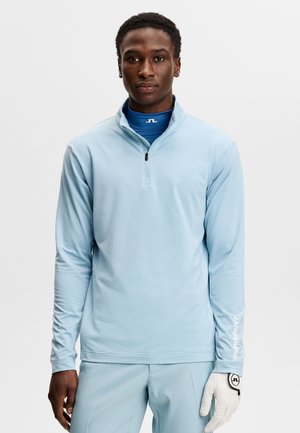 Mann trägt hellblauen Pullover mit Viertel-Reißverschluss und Hose, trägt einen weißen Golfhandschuh an der rechten Hand, steht vor einem schlichten weißen Hintergrund.