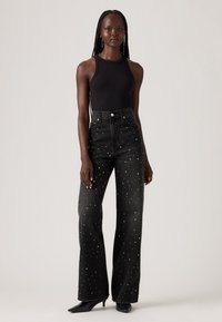 Zwarte high-waisted jeans met witte en zilveren studs, gecombineerd met een zwarte mouwloze top en zwarte haklaarzen.