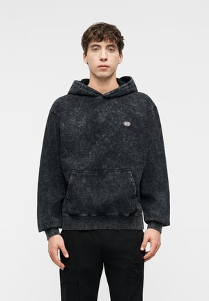 Młody mężczyzna stojący przodem, ubrany w czarną, marmurkową bluzę z kapturem i kieszenią z przodu oraz czarne spodnie, na jednolitym tle.