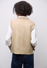 Beige gebreide vest met ribbelranden, gedragen over een witte lange mouwen shirt. Het ontwerp heeft een hoge kraag en een losse pasvorm.