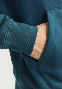 Türkise Sweatshirt mit gerippten Bündchen und einer Kängurutasche. Der Stoff wirkt weich mit einer glatten Textur. Sichtbare Nähdetails.