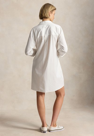 Kvinne med kort blondt hår iført en hvit langermet skjortekjole og hvite joggesko, stående mot en ensfarget beige bakgrunn.