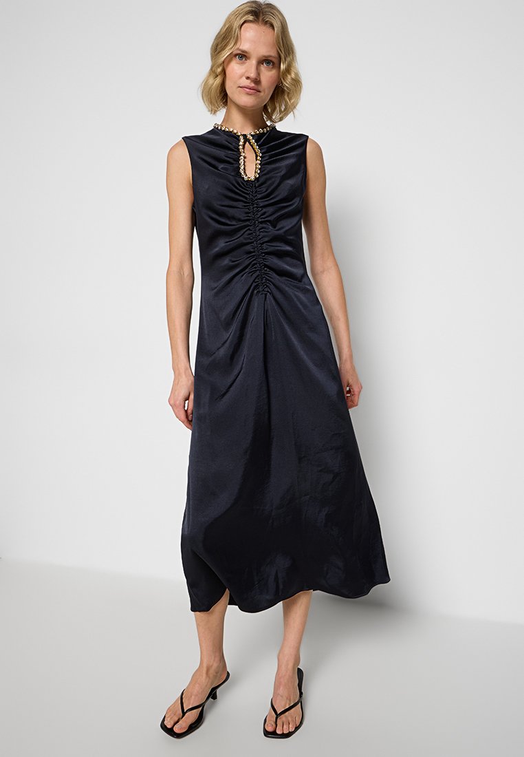 Blonde Frau in einem ärmellosen, gerafften, marineblauen Midikleid mit perlenbesetztem Schlüssel-Ausschnitt und schwarzen, hohen Sandalen, steht vor einer weißen Wand.