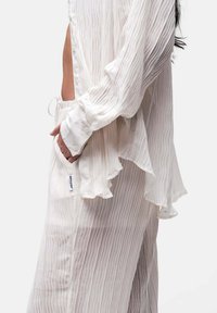 Weiße, plissierte Bluse mit lässigem Schnitt, versehen mit sanften Rüschen am Saum und an den Ärmelbündchen sowie seitlichen Taschen. Subtile Textur sorgt für Tiefe.