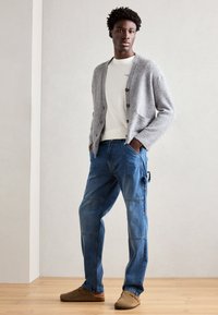 Grå strikket cardigan med knapper, hvit t-skjorte under, blå cargo bukser, og brune klippans på et tregulv mot en nøytral bakgrunn.