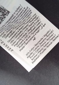 Etichetta con testo multilingue, contenuto del tessuto: 50% cotone, con lingue riportate in testo nero su sfondo bianco.