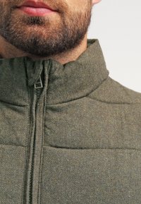 Grüne gesteppte Jacke mit hohem Kragen und Reißverschluss. Hat einen strukturierten Stoff mit dezentem Mélange-Muster und gepolsterten Bereichen.