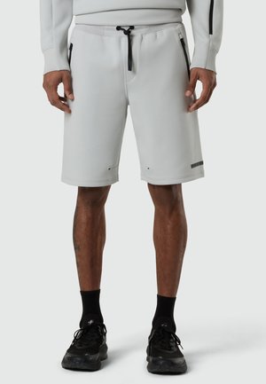 Hombre con pantalones cortos de cordón gris claro, calcetines negros hasta el tobillo y zapatos deportivos negros, de pie con las manos relajadas a los lados.