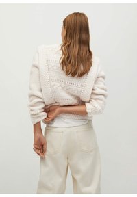 Cardigan en tricot blanc avec des motifs texturés, coupe ample et manches retroussées. Associé à un jean beige taille haute. Vue de dos.