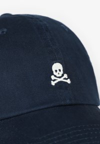 Navy baseball cap with a white embroidered skull and crossbones design on the front, made from textured fabric with stitched detailing.