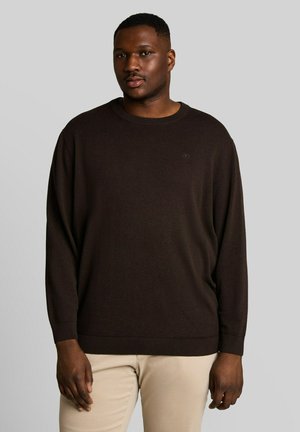 Homme portant un pull marron foncé à manches longues et un pantalon beige, debout devant un fond gris uni.