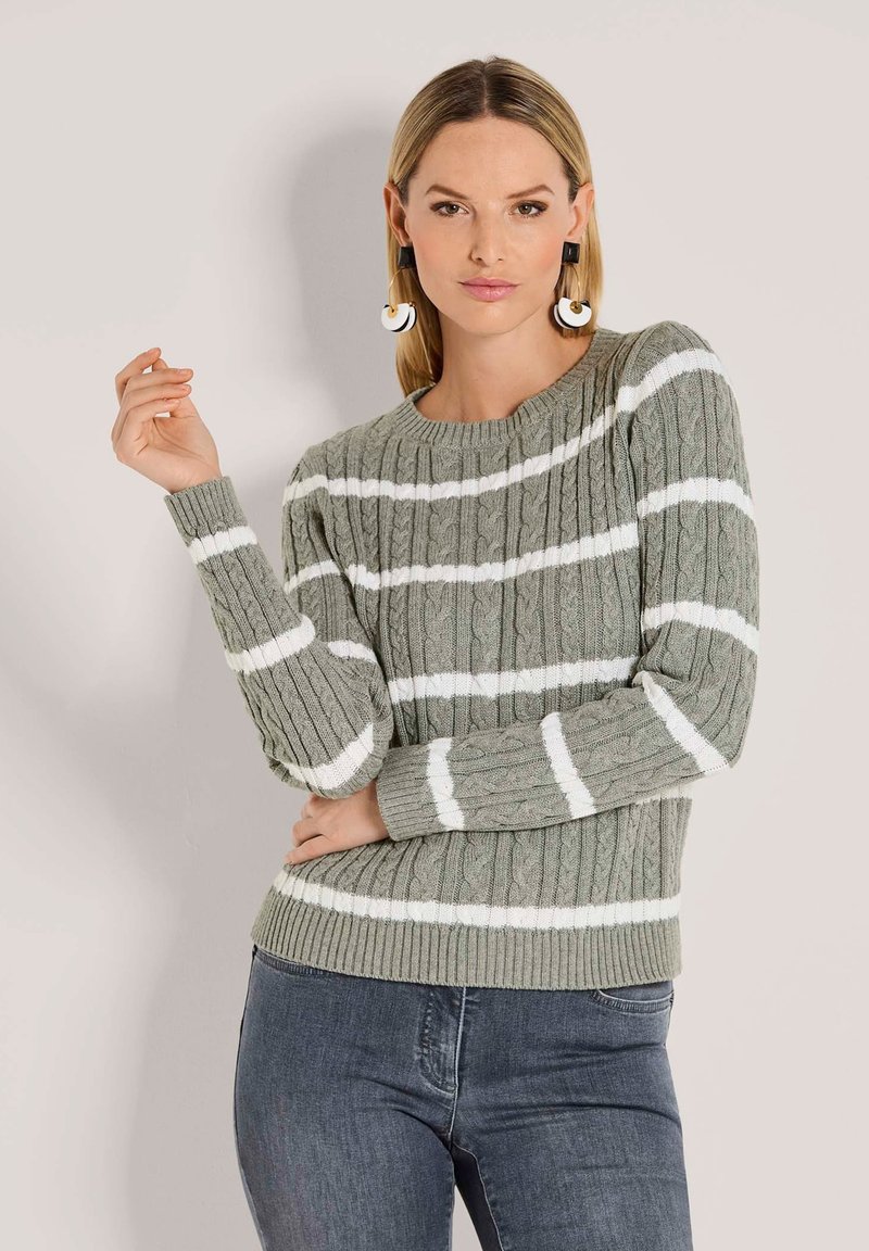 Grauer, kabelgestrickter Pullover mit weißen Streifen, gerippten Bündchen und Saum. Das Model trägt den Pullover mit dunklen Jeans und auffälligen Ohrringen.