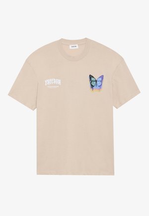 Beige Baumwoll-T-Shirt mit einem kleinen Grafikmotiv eines blauen Schmetterlings mit schwarzen Akzenten und einem Tropfenmuster, sowie weißem Text auf der linken Seite.