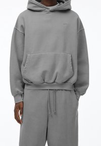 Sweat à capuche gris en tissu doux, avec une poche avant, une capuche à cordon de serrage et des poignets côtelés. Associé à un pantalon de survêtement assorti.