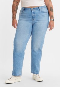 Jeans de denim azul claro con cintura alta, piernas rectas y bolsillos frontales, combinados con zapatos blancos. Tejido liso y texturizado.