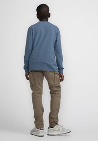 Langemouwen blauwe shirt, kaki cargobroek met zijzakken, en witte schoenen met grijze en gouden accenten. Eenvoudige, casual outfit van achteren.