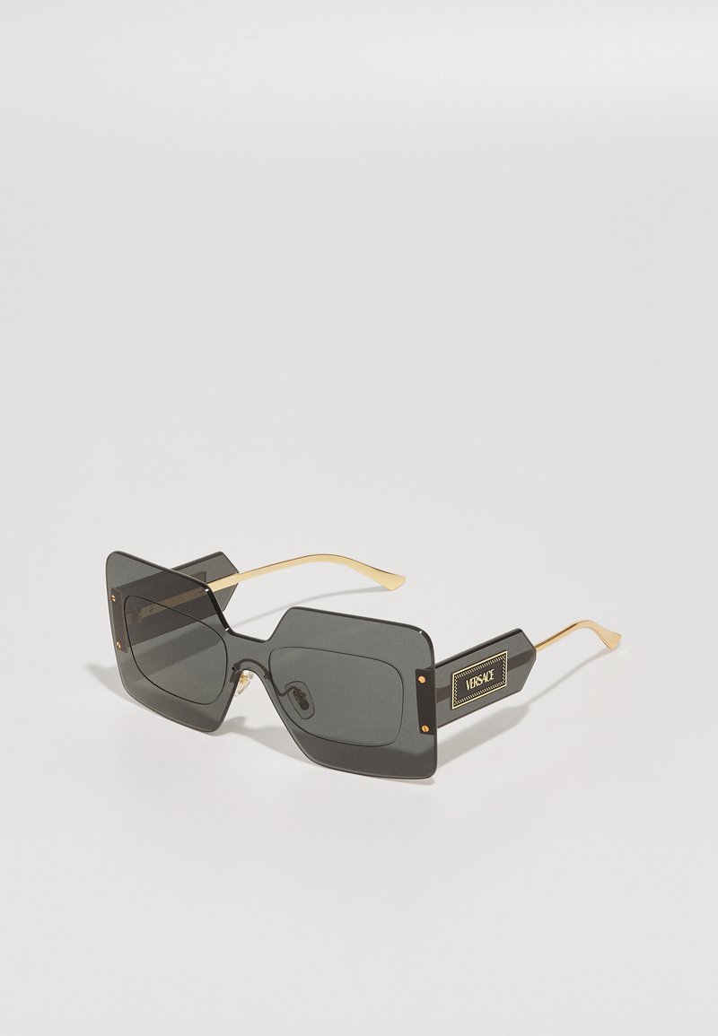 Lunettes de soleil carrées noires avec des accents en métal doré et un logo, présentant des montures épaisses et des verres surdimensionnés. Design en plastique texturé.