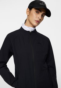 Svart lättviktsjacka med hög krage, dragkedjestängning och broderad logotyp. Slät textur, figurnära design och ventilerande sidopaneler.