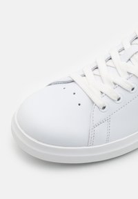 Baskets en cuir blanc avec un bout arrondi, des lacets plats blancs et de petits trous de ventilation. Semelle en caoutchouc blanc texturée. Design minimaliste.