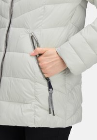 Jasnoszara kurtka ocieplana o pikowanej teksturze, z zamkniętą kieszenią na zamek błyskawiczny i kontrastującymi czarnymi elementami.