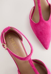Rózsaszín színű, velúr anyagú magassarkú cipő hegyes orral, bokapánttal és arany színű márkajelzéssel a belsejében. Simában tapintású, strukturált kialakítású.