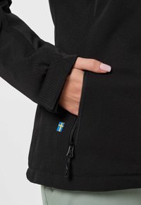 Hand met bleekroze nagels in de met rits afsluitbare zijzak van een zwarte jas met een klein patch van de Zweedse vlag.