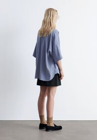 Loose-fitting, short-sleeved blue top with a rounded hem, paired with a black skirt, tan ribbed socks, and black shoes. Smooth fabric texture.