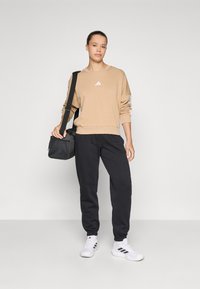 Beige genser med hvitt logo, svarte bukser og hvite joggesko, holder en liten svart veske. Enkelt, avslappet snitt og farger.