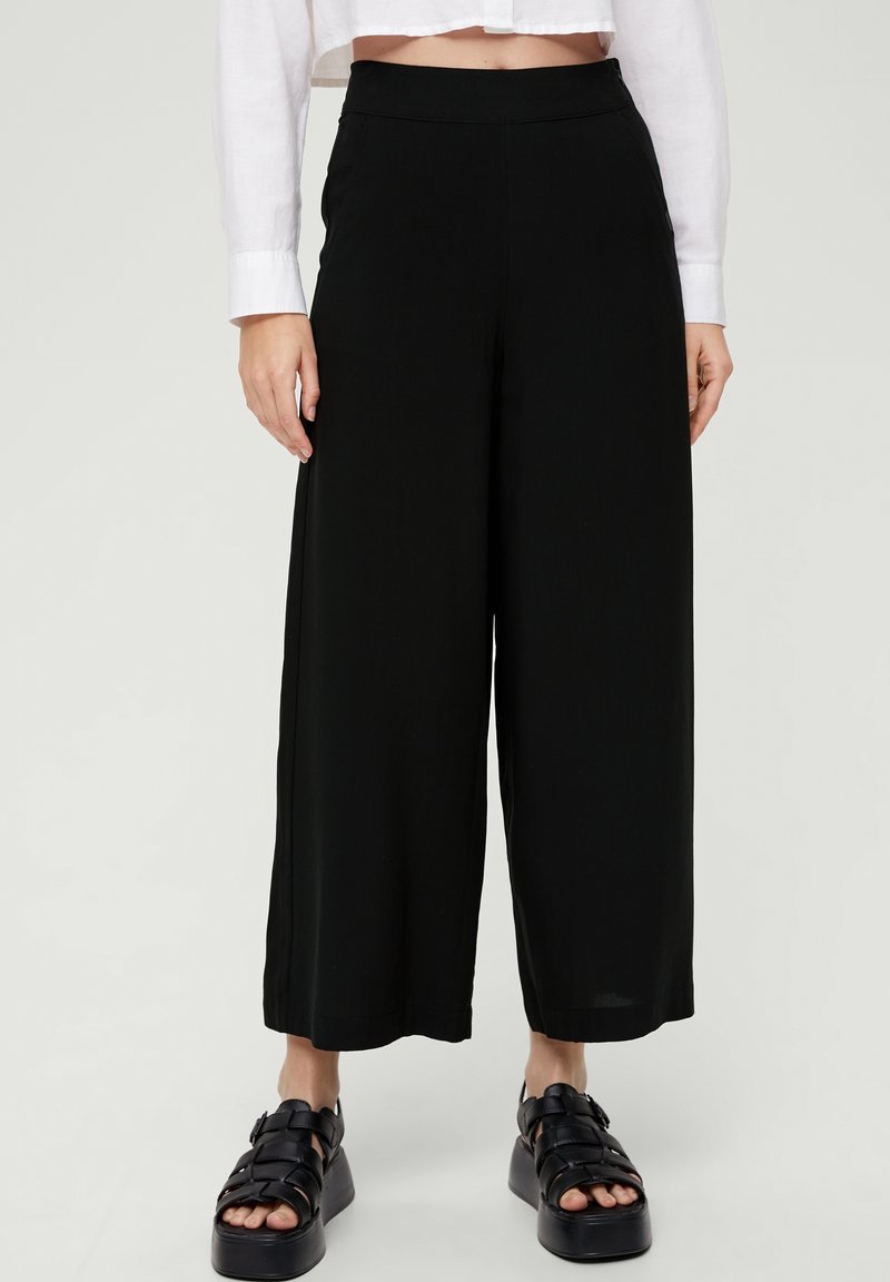 Personne portant un pantalon large noir coupé au-dessus de la cheville, une chemise blanche à manches longues raccourcie et des sandales plateformes noires sur un fond uni.