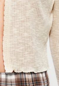 Haut beige en maille côtelée à manches longues associé à un pantalon à carreaux marron et blanc, montrant un gros plan de la zone latérale du torse.