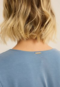 Niebieska bluzka z krótkim rękawem, z okrągłym dekoltem i teksturowanym materiałem. Posiada małą metalową metkę z tyłu.