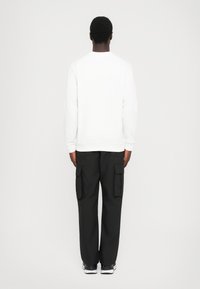 Uomo di spalle che indossa una camicia bianca a maniche lunghe, pantaloni cargo neri con ampie tasche laterali e sneaker bianco e nere.