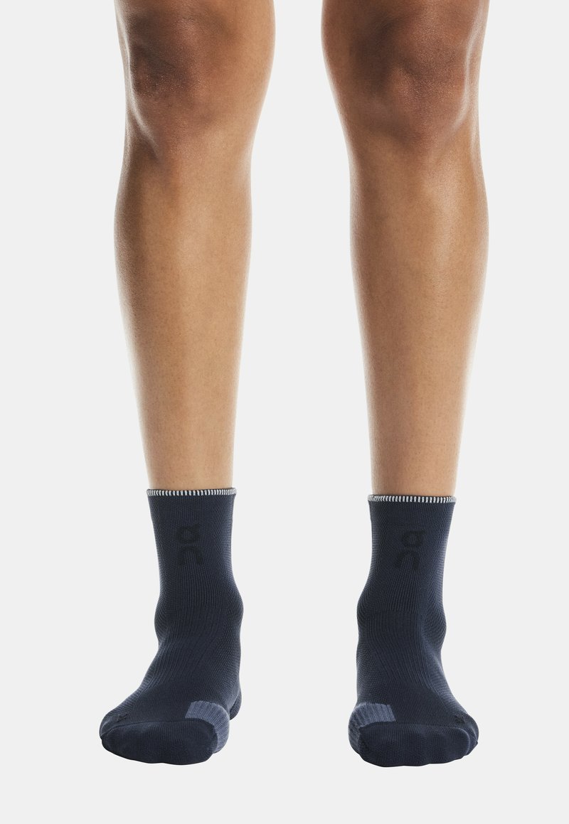 Jambes portant des chaussettes noires courtes avec des semelles texturées antidérapantes, debout sur un fond blanc uni.