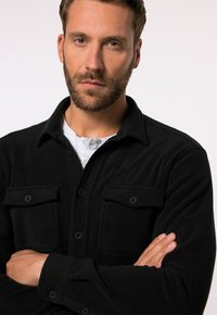 Camisa de forro polar negra con un cierre de botones en la parte delantera, dos bolsillos en el pecho y mangas largas. Textura suave con un cuello tipo solapa.